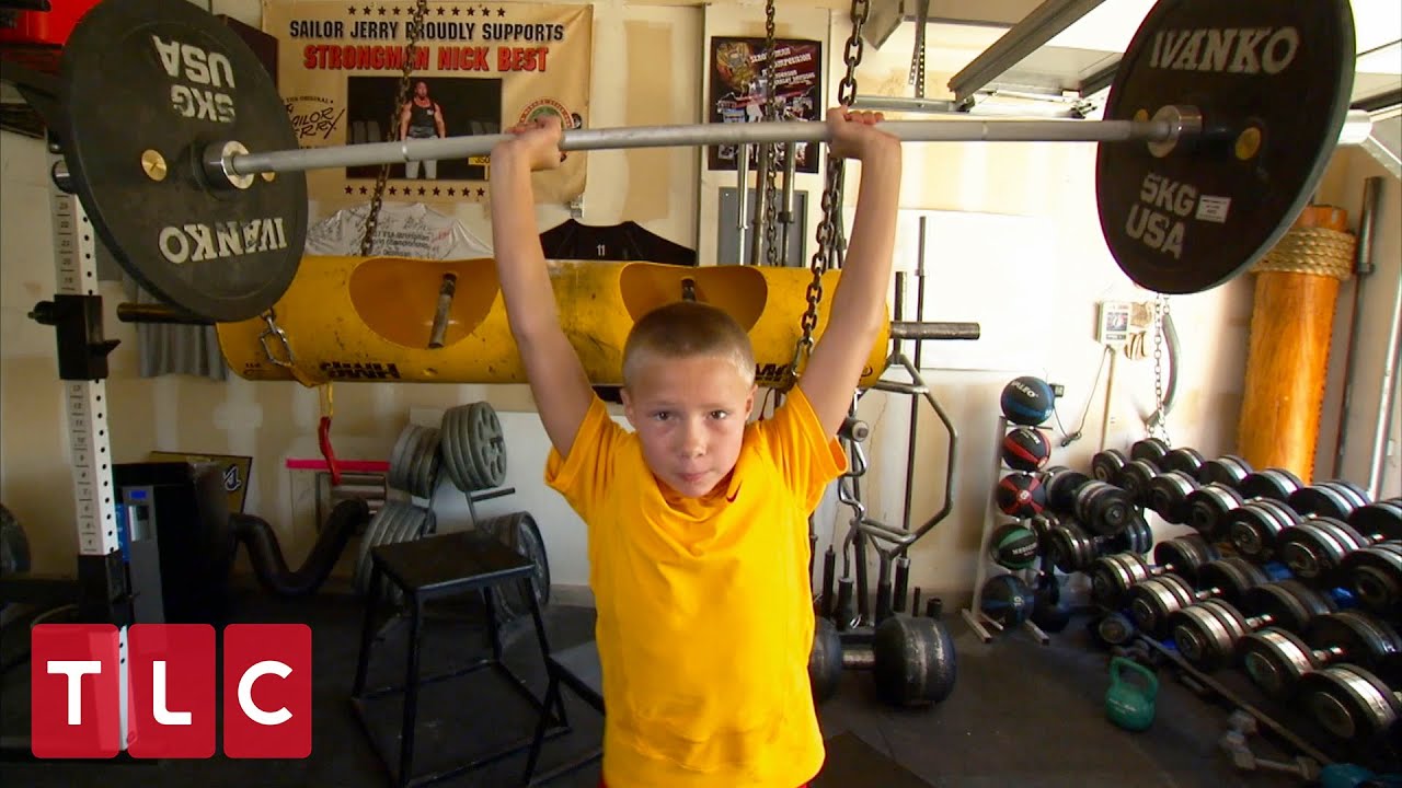 Nick Best’s young son overhead-pressing a barbell beneath a Strongman Nick Best banner